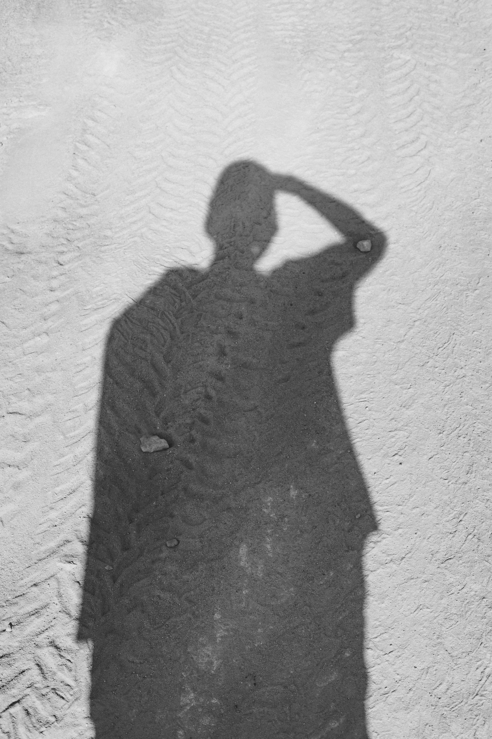 A shadow silhouette of a person cast on a textured sandy beach surface in sunlight.